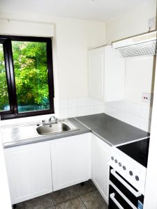 A compact kitchen with white cabinetry and countertops. A sink is beneath a window with a view of green foliage and glimpses of the historic Ruth House. A stove with an extractor fan is to the right. The backsplash is tiled in white, and the floor has gray tiles. Available for sale by auction with Palace Auctions.