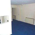 A room with off-white walls and a blue carpeted floor. The room features a closed white door, an electric wall heater, and a small open window-like space revealing part of a kitchen with a stove visible through it. The ceiling has a single exposed bulb. Views of Ruth House available for sale by auction with Palace Auctions.