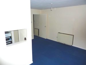 A room with off-white walls and a blue carpeted floor. The room features a closed white door, an electric wall heater, and a small open window-like space revealing part of a kitchen with a stove visible through it. The ceiling has a single exposed bulb. Views of Ruth House available for sale by auction with Palace Auctions.