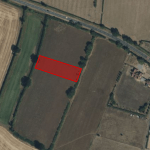 Aerial view of a rural landscape with fields and a red rectangular area marked near the center. The marked parcel of land available for sale at Tilbrook near Huntingdon has trees surrounding it, and there is a road running horizontally near the top. Buildings can be seen near the upper right corner.