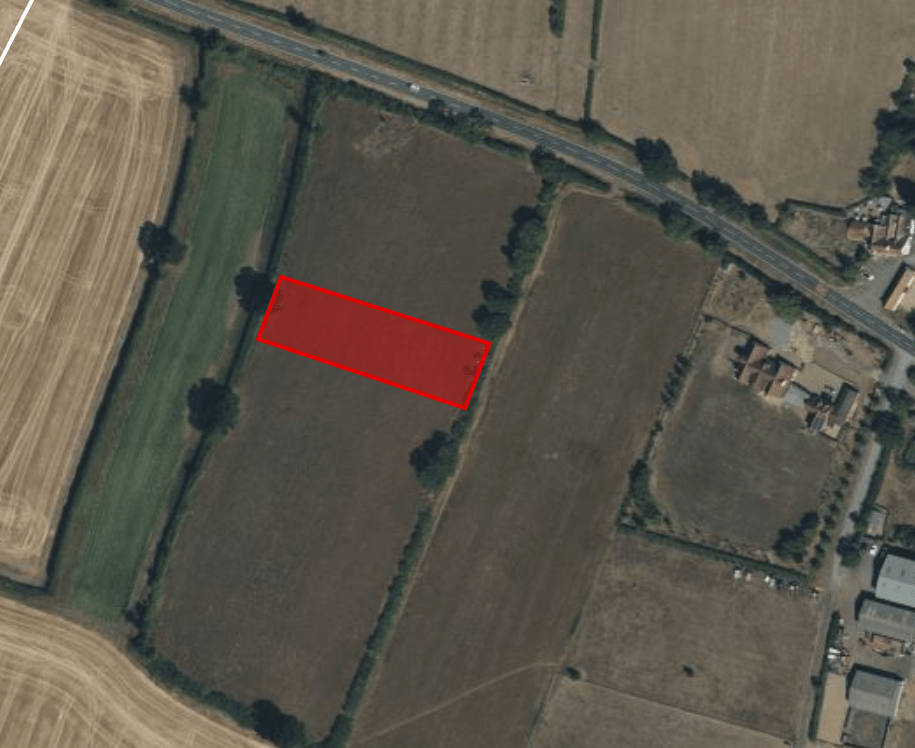 Aerial view of a rural landscape with fields and a red rectangular area marked near the center. The marked parcel of land available for sale at Tilbrook near Huntingdon has trees surrounding it, and there is a road running horizontally near the top. Buildings can be seen near the upper right corner.