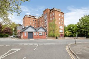 A large brick building with several stories on a corner lot, featuring a modern extension with a sloping roof. Surrounded by trees and greenery, New Alexandra Court Nottingham offers investment apartments for sale by auction through Palace Auctions. The foreground shows road markings and a crosswalk on a clear, sunny day.
