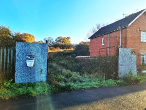 A vacant lot overgrown with grass and weeds is bordered by a red brick building. The lot, a potential development site at the rear of 227-257 Lea Road Gainsborough, is enclosed by a metal fence with two large, gray panels serving as a gate. A poster for an auction with Palace Auctions is attached to one panel. Bright sunlight illuminates the scene.