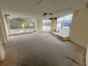 An empty, run-down storefront with large windows on 1-11 Wellington Street, Millom, Cumbria. The ceiling has a fan and fluorescent lights. The floor is covered with debris and the walls are peeling. This historic former co-op building offers a unique residential and commercial conversion opportunity for sale by auction with Palace Auctions.