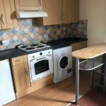 A compact kitchen with wooden cabinets, a patterned tile backsplash, and light wood flooring is featured in investment apartments for sale by auction. It includes a white oven with an electric stovetop, a washing machine, and a small refrigerator. In the foreground, there's a wooden breakfast bar with metal legs.