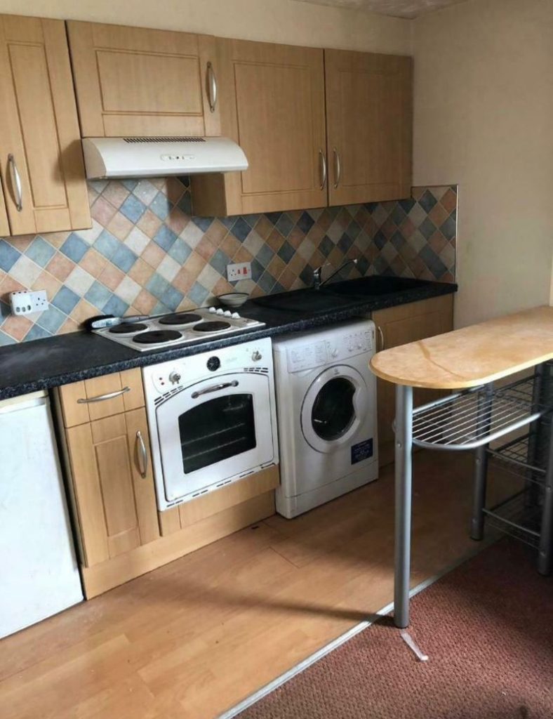 A compact kitchen with wooden cabinets, a patterned tile backsplash, and light wood flooring is featured in investment apartments for sale by auction. It includes a white oven with an electric stovetop, a washing machine, and a small refrigerator. In the foreground, there's a wooden breakfast bar with metal legs.