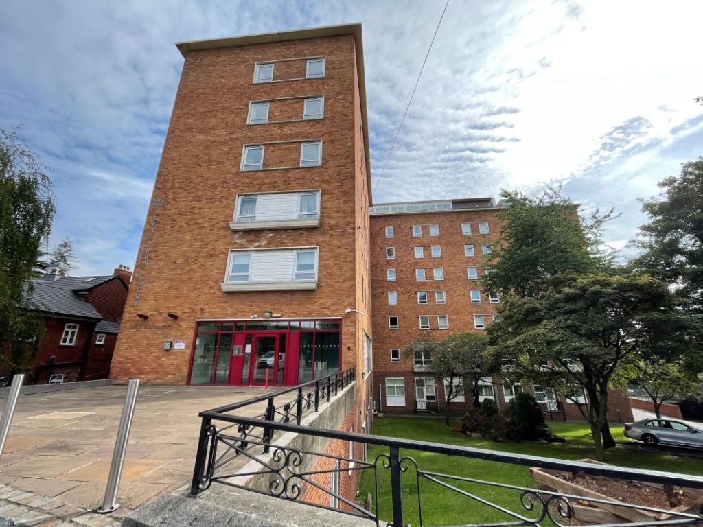 A red-brick, multi-story building with large windows and red-framed entrances. The building is surrounded by a neatly landscaped area with trees and greenery. A black metal railing runs along the foreground, and the sky is partly cloudy.