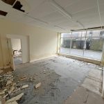 An abandoned, run-down room with broken ceiling tiles and debris scattered across the floor. The walls show signs of water damage and peeling paint. A large window at the far end reveals an empty, wet street outside. Located at 1-11 Wellington Street, Millom, Cumbria, this historic former co-op building offers a residential and commercial conversion development opportunity for sale by auction with Palace Auctions