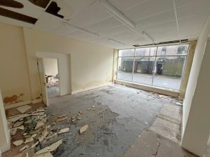 An abandoned, run-down room with broken ceiling tiles and debris scattered across the floor. The walls show signs of water damage and peeling paint. A large window at the far end reveals an empty, wet street outside. Located at 1-11 Wellington Street, Millom, Cumbria, this historic former co-op building offers a residential and commercial conversion development opportunity for sale by auction with Palace Auctions