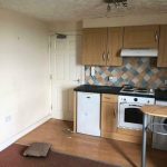 A small kitchen with wooden cabinets, a white oven, and a hood highlights this investment apartment for sale by auction at New Alexandra Court Nottingham. The backsplash features a colorful diamond pattern. A table with a metal leg is partially visible, accompanied by a beige door and an under-counter fridge.