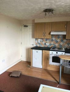 A small kitchen with wooden cabinets, a white oven, and a hood highlights this investment apartment for sale by auction at New Alexandra Court Nottingham. The backsplash features a colorful diamond pattern. A table with a metal leg is partially visible, accompanied by a beige door and an under-counter fridge.