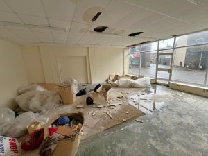 Empty room at 1-11 Wellington Street Millom, Cumbria, with a damaged ceiling, various debris, cardboard boxes, and large trash bags scattered on the floor. Large glass windows allow natural light in and offer a view of the street outside with neighboring buildings. Historic former co-op building - residential and commercial conversion development opportunity for sale by auction with Palace Auctions.