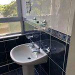A bathroom sink with a blue and white tile backsplash. Above the sink, there is a small shelf with a mirror and a toothbrush holder mounted on the wall. A large window next to the sink offers a view of greenery outside. The floor is tiled in matching blue, complementing the quality in New Alexandra Court Nottingham's investment apartments for sale by auction.