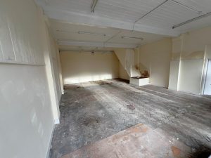 An empty room at 1-11 Wellington Street Millom, Cumbria, with cream-colored walls and a high white ceiling. The floor is made of dark, herringbone-patterned wooden planks. White ceiling lights illuminate areas where paint appears to be peeling. Natural light fills this historic former co-op building—an ideal residential and commercial conversion development opportunity for sale by auction with Palace Auctions