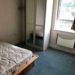 A small, sparsely furnished bedroom featuring a single bed with an unmade mattress and a wooden frame, a white wardrobe with mirrored doors, a light blue carpet, a window with partially pulled back curtains, and a wall-mounted heater. Ideal for those looking at investment apartments for sale by auction.