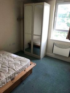 A small, sparsely furnished bedroom featuring a single bed with an unmade mattress and a wooden frame, a white wardrobe with mirrored doors, a light blue carpet, a window with partially pulled back curtains, and a wall-mounted heater. Ideal for those looking at investment apartments for sale by auction.