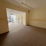 A vacant room at 1-11 Wellington Street, Millom, Cumbria, part of the historic former co-op building. This residential and commercial conversion development opportunity features beige walls and brown carpet flooring. Extending into another area via a doorway, large windows on the far side allow natural light to enter. Some cables and debris are visible on the floor. For sale by auction with Palace Auctions