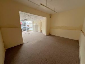 A vacant room at 1-11 Wellington Street, Millom, Cumbria, part of the historic former co-op building. This residential and commercial conversion development opportunity features beige walls and brown carpet flooring. Extending into another area via a doorway, large windows on the far side allow natural light to enter. Some cables and debris are visible on the floor. For sale by auction with Palace Auctions