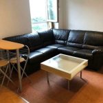 A cozy living room at New Alexandra Court Nottingham features a black leather L-shaped sofa positioned in the corner. In front of the sofa is a white square coffee table. An open window allows natural light to illuminate the space. Discover investment apartments for sale by auction with Palace Auctions.