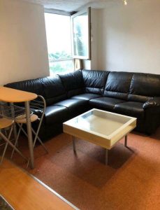 A cozy living room at New Alexandra Court Nottingham features a black leather L-shaped sofa positioned in the corner. In front of the sofa is a white square coffee table. An open window allows natural light to illuminate the space. Discover investment apartments for sale by auction with Palace Auctions.