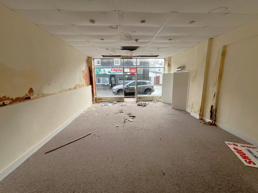 An empty, run-down room with a damaged ceiling, peeling paint, and scattered debris on the carpeted floor lies within the historic former co-op building at 1-11 Wellington Street Millom Cumbria. The glass front window shows a view of parked cars on the street. Walls display signs of water damage and mold growth.