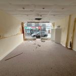 An empty, run-down room with a damaged ceiling, peeling paint, and scattered debris on the carpeted floor lies within the historic former co-op building at 1-11 Wellington Street Millom Cumbria. The glass front window shows a view of parked cars on the street. Walls display signs of water damage and mold growth.
