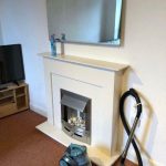 A living room in an investment apartment for sale by auction at New Alexandra Court, Nottingham, features a white fireplace with a reflective mirror above it and two cleaning spray bottles on its mantle. Below, a modern fireplace insert displays white decorative stones. A blue vacuum cleaner rests in front of the fireplace. A flat-screen TV on a wooden stand is visible to the left. The room has beige carpet and white