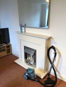 A living room in an investment apartment for sale by auction at New Alexandra Court, Nottingham, features a white fireplace with a reflective mirror above it and two cleaning spray bottles on its mantle. Below, a modern fireplace insert displays white decorative stones. A blue vacuum cleaner rests in front of the fireplace. A flat-screen TV on a wooden stand is visible to the left. The room has beige carpet and white