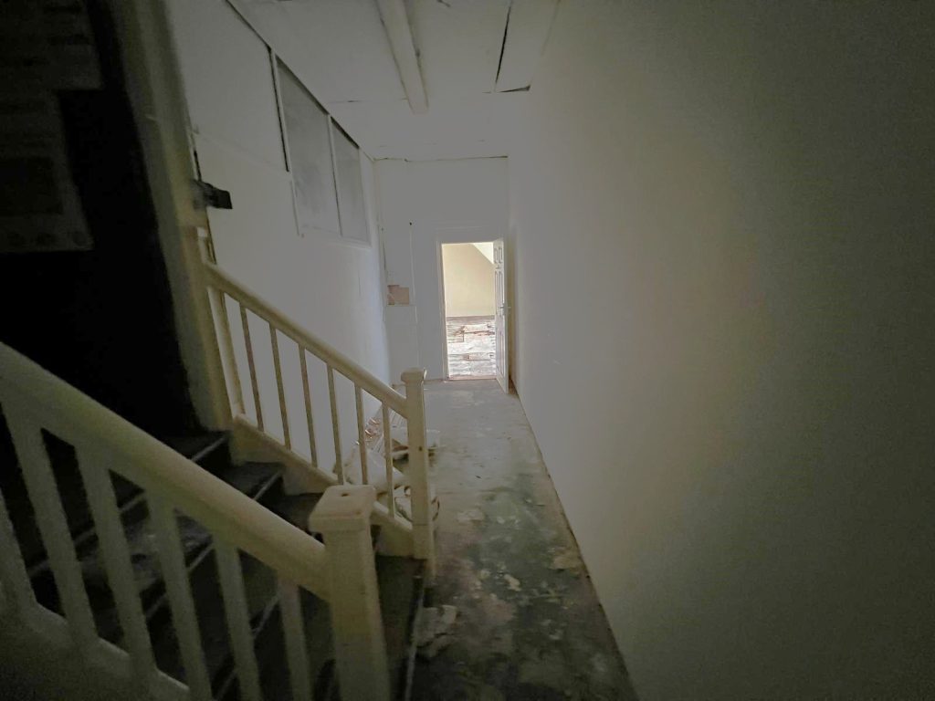 A dimly lit hallway with peeling paint on the walls and a bare floor leads through 1-11 Wellington Street, Millom, Cumbria. On the left, there's a staircase with white railings leading upwards. At the end of this historic former co-op building's hallway, a doorway opens to a sunlit area with debris scattered on the ground.