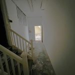 A dimly lit hallway with peeling paint on the walls and a bare floor leads through 1-11 Wellington Street, Millom, Cumbria. On the left, there's a staircase with white railings leading upwards. At the end of this historic former co-op building's hallway, a doorway opens to a sunlit area with debris scattered on the ground.