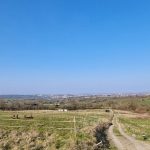 A wide landscape view of a countryside with grassy fields, a dirt path winding through the right, and scattered trees. In the distance, a town under a clear blue sky can be seen. The scenery includes gentle rolling hills and open spaces enclosed by fences, reminiscent of PLOT C38 DUNDRY LANE Bristol sold by Palace Auctions.