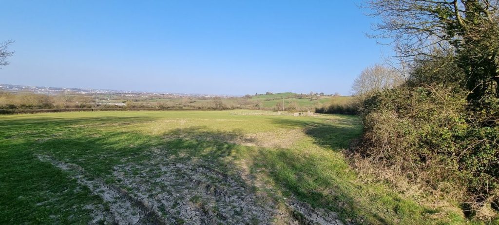 A wide, grassy field extends toward distant, rolling hills under a clear, blue sky. Trees and bushes frame the field on the right side, while buildings can be seen far off on the horizon. The ground is a mix of green grass and bare patches of earth, reminiscent of plot C38 Dundry Lane Bristol sold by Palace Auctions.