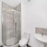 A modern bathroom in the Humphry Davy House investment flat available for sale by auction features a glass-enclosed corner shower with grey tiled walls, a white toilet, and a white sink with a grey tile backsplash. Predominantly white, the room includes a silver towel radiator on the left side.