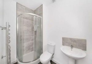 A modern bathroom in the Humphry Davy House investment flat available for sale by auction features a glass-enclosed corner shower with grey tiled walls, a white toilet, and a white sink with a grey tile backsplash. Predominantly white, the room includes a silver towel radiator on the left side.