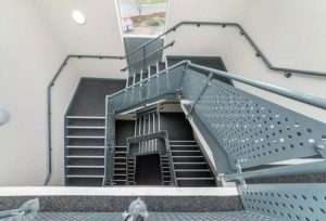 A modern multi-story stairwell with grey perforated metal railings and dark carpeting on the steps. The stairwell features several flights of stairs and landings, winding down to the building's lower floors. A window at the top lets in natural light, enhancing this Humphry Davy House investment flat available for sale by auction.