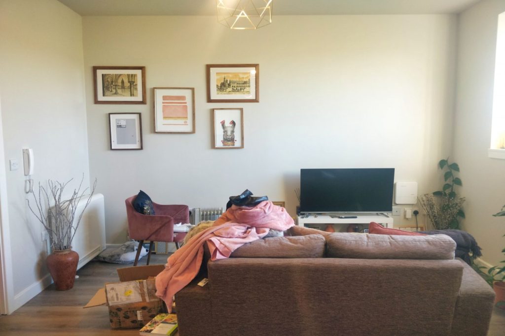 A cozy living room with a brown couch facing a flat-screen TV on a console. An armchair is partially covered with blankets, and various items and plants are scattered around. Four framed artworks decorate the wall above the TV in this inviting space, now available for sale by auction at Humphry Davy House. Sunlight streams in through a window.