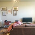 A cozy living room with a brown couch facing a flat-screen TV on a console. An armchair is partially covered with blankets, and various items and plants are scattered around. Four framed artworks decorate the wall above the TV in this inviting space, now available for sale by auction at Humphry Davy House. Sunlight streams in through a window.
