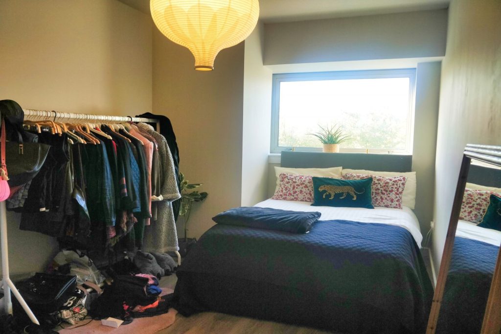 A small bedroom in the Humphry Davy House investment flat, available for sale by auction with Palace Auctions, features a double bed adorned with blue and white bedding and a decorative leopard pillow. A clothing rack filled with various garments stands to the left, while natural light streams in through a large window.