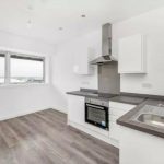 A modern kitchen in the Humphry Davy House investment flat is available for sale by auction through Palace Auctions below market value. It features white cabinets, grey countertops, a built-in oven, a stovetop with a stainless steel hood, and large windows offering an ocean view.