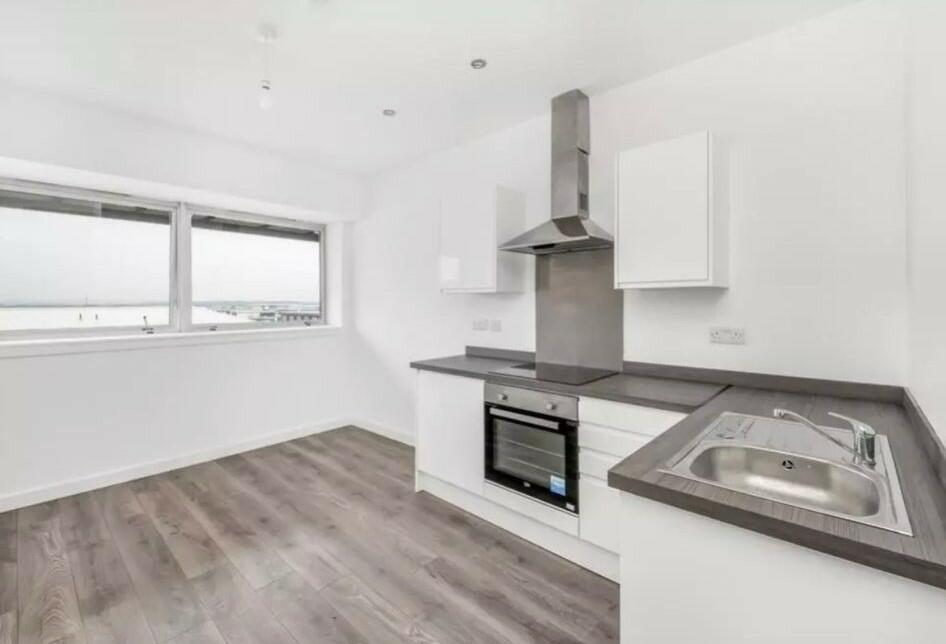 A modern kitchen in the Humphry Davy House investment flat is available for sale by auction through Palace Auctions below market value. It features white cabinets, grey countertops, a built-in oven, a stovetop with a stainless steel hood, and large windows offering an ocean view.