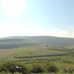 A lush green landscape featuring rolling hills under a clear, blue sky. The foreground has dense vegetation with various shades of green, while the background shows expansive grassy hills and a few scattered clouds. This serene scene mirrors the beauty of Land off Channel Avenue, Porth, Mid Glamorgan, Wales (SOLD) by Palace Auctions.