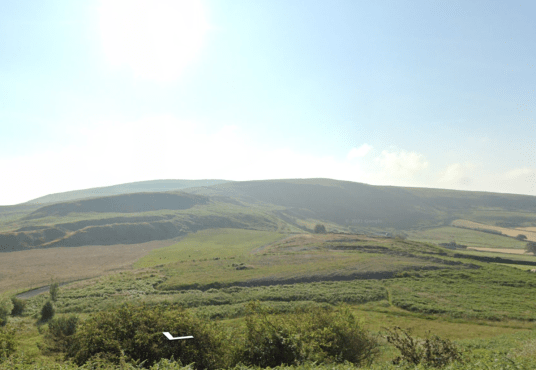 A lush green landscape featuring rolling hills under a clear, blue sky. The foreground has dense vegetation with various shades of green, while the background shows expansive grassy hills and a few scattered clouds. This serene scene mirrors the beauty of Land off Channel Avenue, Porth, Mid Glamorgan, Wales (SOLD) by Palace Auctions.