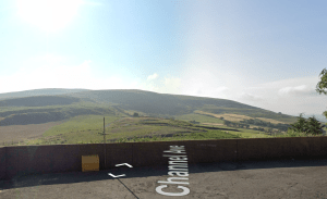 A scenic view of rolling green hills under a clear blue sky. In the foreground, part of a brick wall with a yellow waste bin can be seen. The road labeled "Channel Ave" runs along the bottom of the image. Land off Channel Avenue Porth Mid Glamorgan Wales (SOLD) by Palace Auctions - other plots may be available; contact the auctioneers.