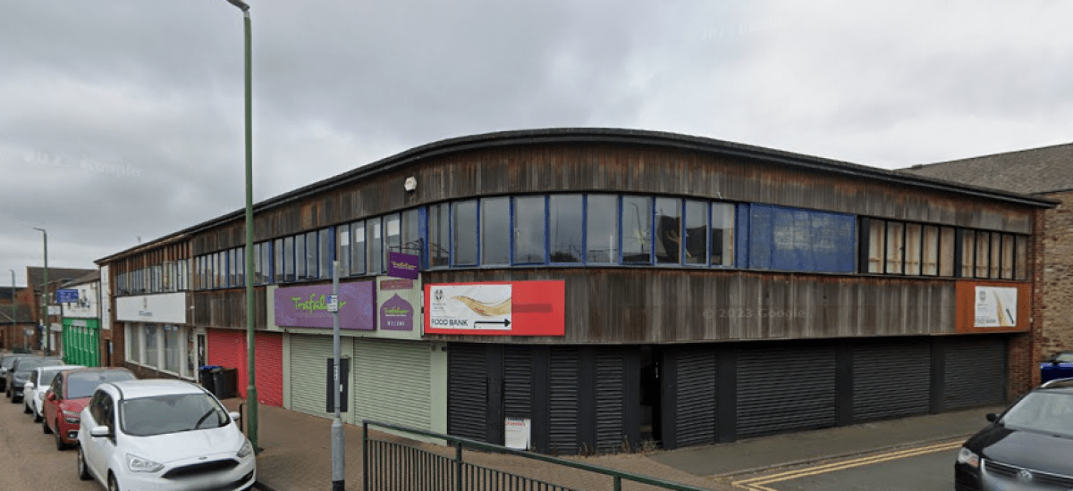A two-story commercial building with wooden cladding and large windows on the upper floor is located on Trafalgar Street, Consett. Several business signs are displayed, and the lower storefronts have their shutters down. Cars are parked along the street under an overcast sky. The commercial investment property was sold by Palace Auctions.