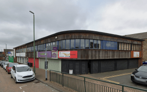 A two-story commercial building with wooden cladding and large windows on the upper floor is located on Trafalgar Street, Consett. Several business signs are displayed, and the lower storefronts have their shutters down. Cars are parked along the street under an overcast sky. The commercial investment property was sold by Palace Auctions.