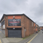 Street view of a red brick, two-story boxing and weights gym on Trafalgar Street, Consett, with a large advertisement above the entrance. The gym is situated on a narrow street with buildings and cars lining the sides. The cloudy sky overhead gives a greyish ambiance.