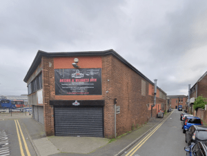 Street view of a red brick, two-story boxing and weights gym on Trafalgar Street, Consett, with a large advertisement above the entrance. The gym is situated on a narrow street with buildings and cars lining the sides. The cloudy sky overhead gives a greyish ambiance.