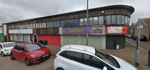 Street view of a two-story commercial investment property on Trafalgar Street, Consett, featuring multiple businesses like the Trafalgar Pub and Eatery and a Food Bank. The building has a curved wooden facade on the upper floor and colorful signage, with cars parked in front. This property was sold by Palace Auctions.