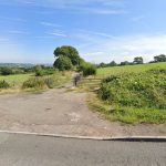 A country road curves alongside a grassy field with a scenic view in the background. The field at Plot C38 Dundry Lane, Bristol is bordered by a wooden gate and lush greenery. Trees and rolling hills extend into the distance under a clear blue sky, sold by Palace Auctions.
