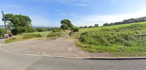 A country road curves alongside a grassy field with a scenic view in the background. The field at Plot C38 Dundry Lane, Bristol is bordered by a wooden gate and lush greenery. Trees and rolling hills extend into the distance under a clear blue sky, sold by Palace Auctions.